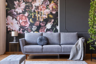 Cushion and blanket on a stylish sofa in a grey living room interior with ivy plant and flowers print on the wall. Maximalist style